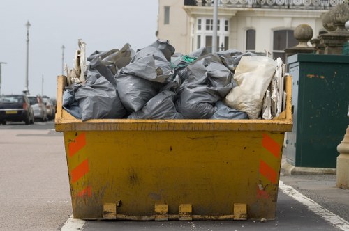 A large yellow skip bin filled with black and white rubbish bags, positioned on a residential street surface in Teddington. The bags appear to contain garden waste, household waste, or debris, and are stacked to the brim, with some bags leaning against each other. The skip shows signs of dirt and weathering, with reflective orange and silver safety markings on its sides. In the background, parked cars are visible along the street, with some building facades partially seen behind a row of decorative stone features and greenery. The scene is outdoors, under natural daylight, indicating an active rubbish removal process that would be handled by services such as Garden Clearance Teddington, specialists in garden and general rubbish clearance services in the local area.
