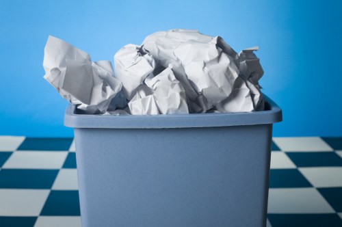 A light blue plastic storage bin filled with crumpled white and gray paper waste, placed on a black and white checkered floor. The background features a smooth, vibrant blue wall, providing a clean and simple setting. The paper waste appears to be typical household or garden rubbish, with various textured crumpled pieces stacked loosely inside the bin. This image reflects the process of gathering waste materials, likely part of a garden clearance service in Teddington, with Garden Clearance Teddington handling rubbish removal tasks. The lighting is even and highlights the crumpled paper's irregular folds and textures, emphasizing the contrast between the smooth plastic bin and the rough, crumpled paper, suitable for illustrating garden or household rubbish collection and disposal themes in a professional context.