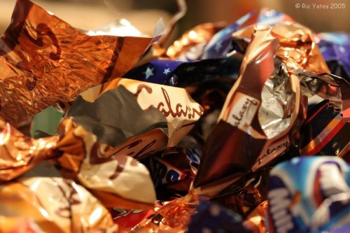 A close-up photograph of a pile of crumpled candy wrappers and foil packaging, predominantly in metallic copper, silver, and shades of blue and white, with some printed branding and decorative patterns visible. The wrappers are scattered and overlapping, creating a chaotic arrangement with textures reflecting light, indicating they are made of thin, reflective foil and paper materials. The background is blurred, suggesting an indoor setting or a container filled with rubbish. The image visually represents waste accumulation typical of rubbish removal situations, though it does not specify a particular environment like a garden or driveway. This scene exemplifies the type of non-organic waste that Garden Clearance Teddington might handle in their rubbish clearance services, especially when dealing with discarded packaging from various products. The image’s focus on the refuse pile emphasizes the importance of proper rubbish disposal and clearance services for preventing clutter and maintaining tidy environments, consistent with the service category for the Teddington area postcodes.