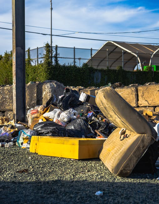 A street scene featuring a waste collection worker loading garbage into a large, red industrial refuse truck parked on the roadside. The worker, dressed in a yellow high-visibility jacket with red acc