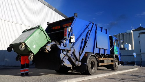 A large blue and black rubbish removal truck with hydraulic arms lifting a green wheeled waste container in an outdoor urban setting, with a modern white industrial building and a clear blue sky in the background. The green waste bin is partially lifted and positioned at the rear of the truck, which is parked on a paved area. A worker wearing high-visibility red and orange clothing is nearby, assisting with the waste collection process. The scene suggests a professional rubbish collection operation, typical of services offered by companies like Garden Clearance Teddington, supporting local waste disposal in the Teddington area and surrounding postcode regions.
