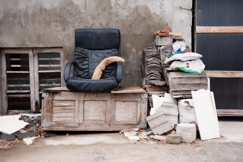 A black office chair with fabric upholstery and metal armrests positioned on a wooden pallet in an outdoor setting. To the right of the chair, there is a pile of construction or building waste, including grey concrete blocks, bricks, and broken pieces of plasterboard, stacked irregularly against a weathered concrete wall. Some debris is scattered on the ground around the pile. The background features a ventilation grille set into the wall and a partly visible black metal storage door with a wooden reinforcement beam. The area appears to be a storage or disposal space in a gritty, urban environment, consistent with rubbish removal services in Teddington or nearby areas, supported by the context of garden clearance or waste collection experts.